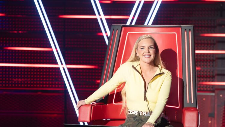 Tones And I seated in a red coach chair on The Voice