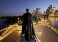 Batman stands above the Sydney Opera House with the illuminated Sydney skyline behind him at dusk.