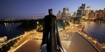 Batman stands above the Sydney Opera House with the illuminated Sydney skyline behind him at dusk.