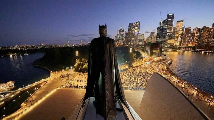 Batman stands above the Sydney Opera House with the illuminated Sydney skyline behind him at dusk.