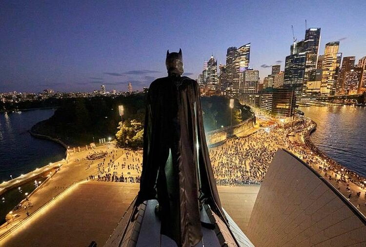 Batman stands above the Sydney Opera House with the illuminated Sydney skyline behind him at dusk.