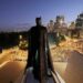 Batman stands above the Sydney Opera House with the illuminated Sydney skyline behind him at dusk.
