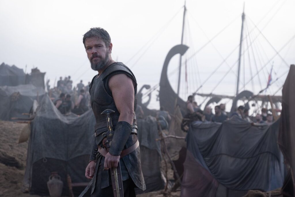 A warrior stands on a beach beside ships in The Odyssey