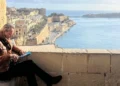 Richard E Grant sits on a stone ledge above a Maltese harbour