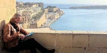 Richard E Grant sits on a stone ledge above a Maltese harbour
