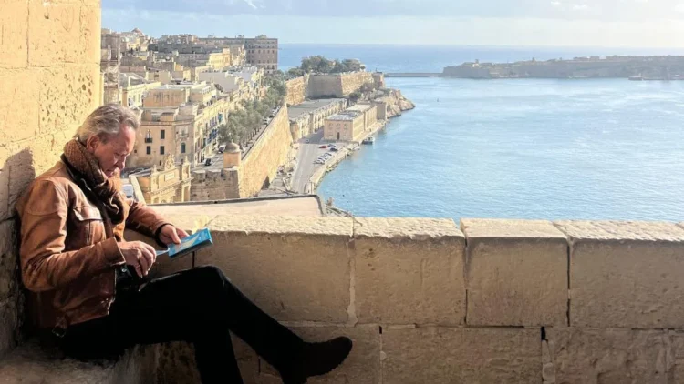 Richard E Grant sits on a stone ledge above a Maltese harbour