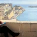 Richard E Grant sits on a stone ledge above a Maltese harbour