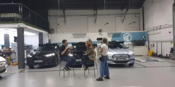 Hannah Fry talks with two people beside cars in a vehicle testing facility