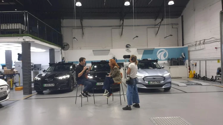 Hannah Fry talks with two people beside cars in a vehicle testing facility