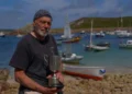 A man on the rocky shore of the Isles of Scilly with boats in the harbour