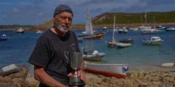 A man on the rocky shore of the Isles of Scilly with boats in the harbour