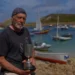 A man on the rocky shore of the Isles of Scilly with boats in the harbour